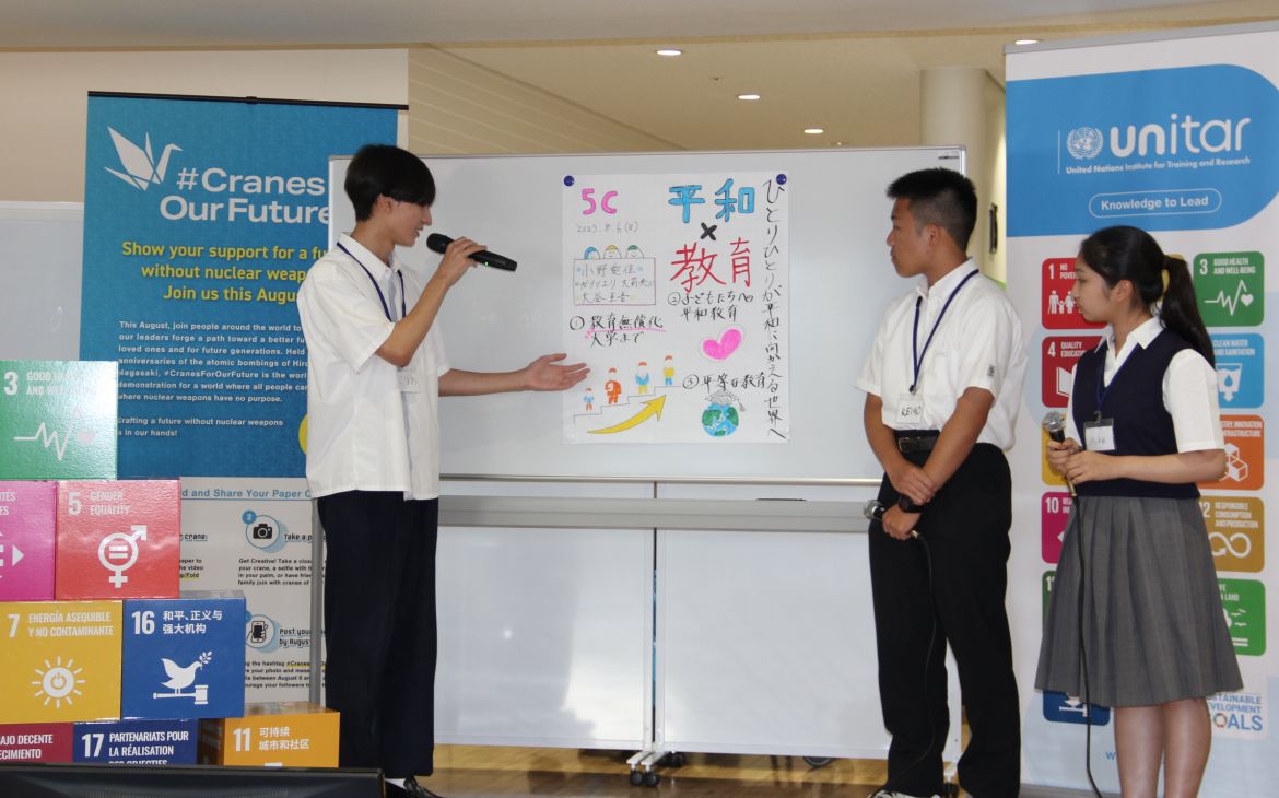 A group of Japanese students consisting of two males and one female presents in front of an audience (not shown). The male student on the left is speaking while the other members standing on the right side are looking on the presentation. The backgound is a whiteboard with a large white paper pasted on it containing some illustrations words written in Japanese while the left and right side of the background consists of the UNITAR logo and the SDG goals logos.