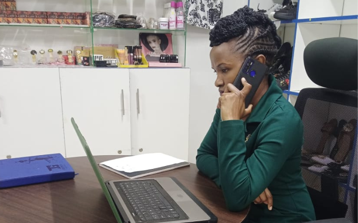 Angeline Mueni on a phone while sitting on her desk facing a laptop at Angekelly store