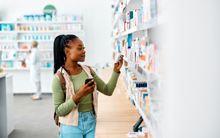 Young African woman in a drugstore choosing a medicine while facing a shelf and with a phone on one hand