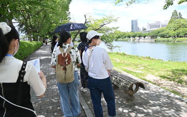 A group of girl students with clipboards walking along a sunny, hot riverbank in Hiroshima. The girls wear caps. One holds an umbrella.