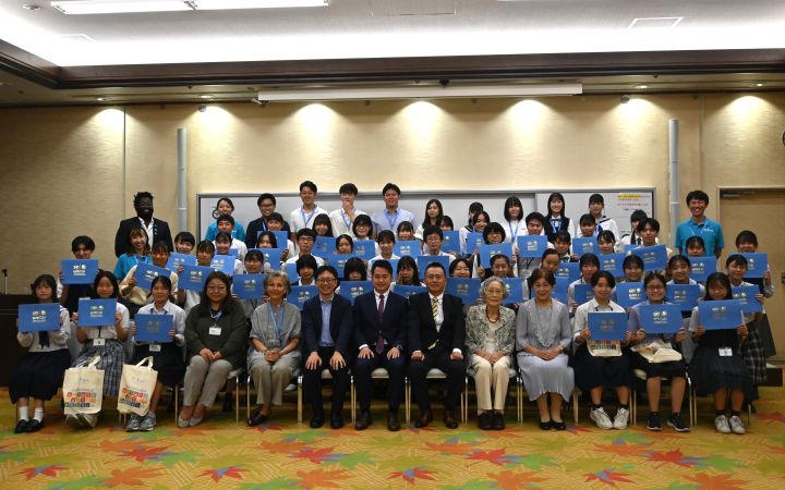 A group photo in a meeting room. Four rows of smiling students all hold up their blue certificates of completion. Seven men and women in business attire sit centre, front row.