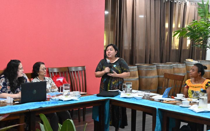 A photo of a Filipina standing (center). She is holding a microphone as she speaks and beside her (left and right) are women from different nationalities. They are seated while listening to the Filipina woman.