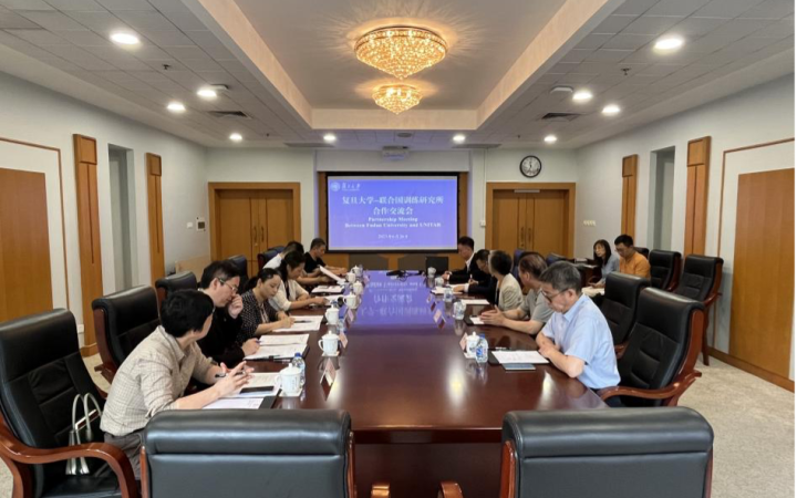 About 15 people are seated in black leather chairs around a long, gleaming table in a carpeted meeting room. Some people are jotting notes on the paper before them, others are looking at the blue slides projected on the screen at the wall at the far end of the table.