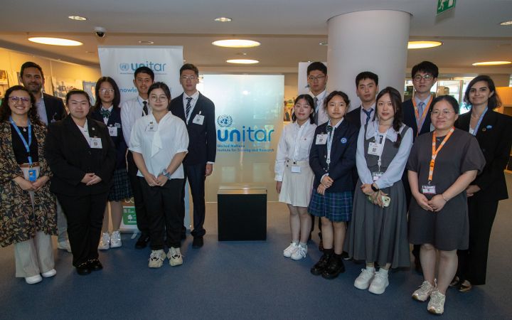 Students, UNITAR representatives, and expert panelist at UNITAR headquarters for the Global Challenges Seminar Group photo of 15 students and professionals, standing in two rows around a glass UNITAR sign in a building lobby