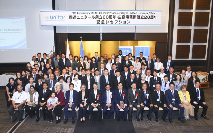 A group photo featuring men and women in their corporate attires. People in the first row of the photo are seating while the rest stands. The main background is a folding panel coated in gold. On the wall hangs a long banner where the logo of UNITAR is shown followed by "60th Anniversary of UNITAR and 20th Anniversary of UNITAR Hiroshima Office".