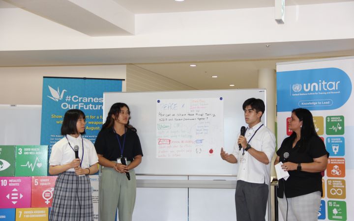 A photo of a group of Japanese students presenting in front of an audience (not shown). On the left side are two female students holding microphones as they listens to the male student speaking on the right. Right beside the male student is what seems to be a event co-facilitator. The backgound is a whiteboard with a large white paper pasted on it containing the keywords from the presenters while the left and right side of the background consists of the UNITAR logo and the SDG logos.