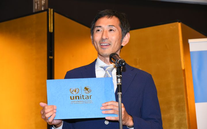 A close-up shot of an Asian man in dark blue suit speaking using a microphone attached to a stand while holding a certificate cover with the logo of UNITAR. The background is a stretch of gold-painted collapsible panels.