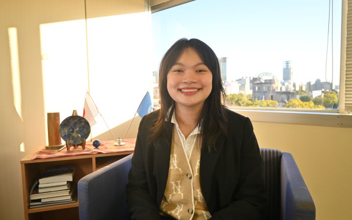 A smiling young Vietnamese woman wearing a black blazer suit as outer wear. She is is sitting on a chair and behind her is a glass window showing the view outside including the Hiroshima Atomic Bomb Dome (right) and on her left a book shelf where small flags of Japan and the United Nations stands forming an X shape.