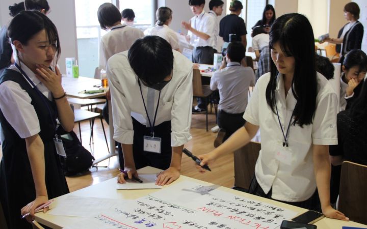 A photo featuring three Japanese (two females and a male between them) students discussing duirng a group activity session. At the bottom of the photo is a large white paper where some Japanese characters are written. The background consists of students doing the same in small tables.