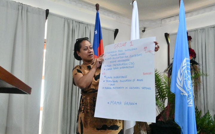 A woman from a Pacific small island developing state stands in front an audience (not shown) as she holds a large piece of white paper containing some texts which she is reading. Behind her are the flags of Samoa and Japan (both partially hidden) and the United Nations flag – all hanging on small flagpoles.