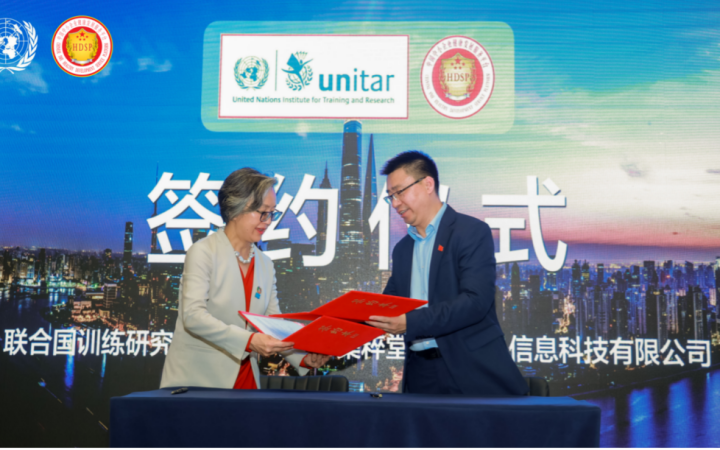 An Asian woman in a suit and glasses and an Asian man with short hair in dark suit exchange two red folders in front of a backdrop with UNITAR and JICUITANG logos and the words “signing ceremony” printed in huge Chinese letters on a photo of Shanghai at sunset.]