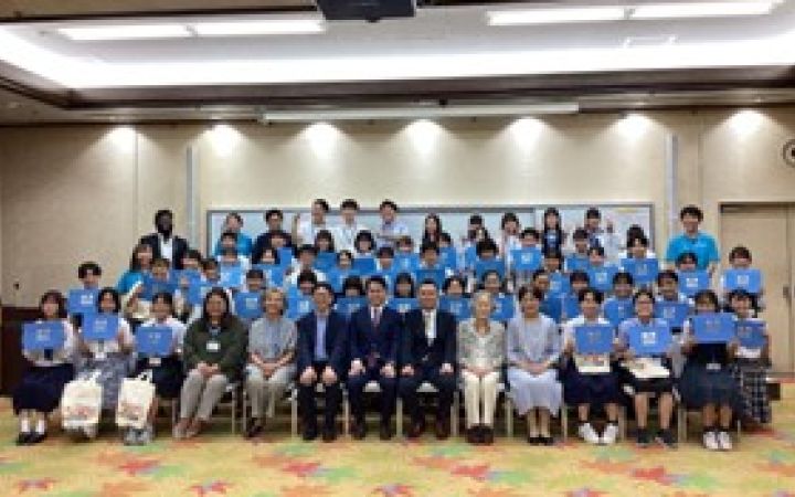 A group photo in a meeting room. Four rows of smiling students all hold up their blue certificates of completion. Seven men and women in business attire sit centre, front row.