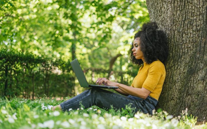 An African woman sitting outdoors while using a laptop with her back resting on a big tree. The background is a scenery of green trees and green grasses at the lower foreground.