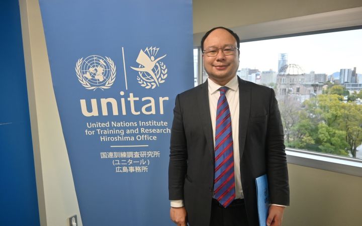 A Malaysian man in black suit and wearing glasses poses for a photo beside UNITAR banner and the Hiroshima skyline featuring the Hiroshima Atomic Bomb Dome as a background on the right side.